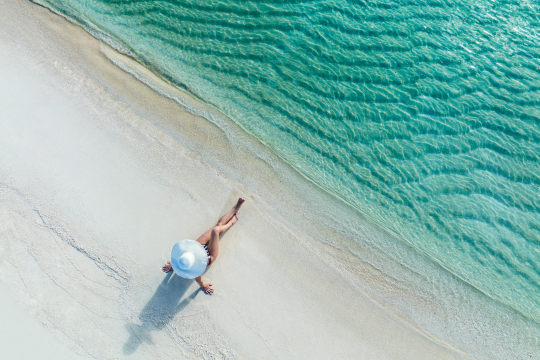Frau mit wei&szlig;em Sonnenhut sitzt an einem wei&szlig;em Sandstrand mit t&uuml;rkisfarbenen Wasser