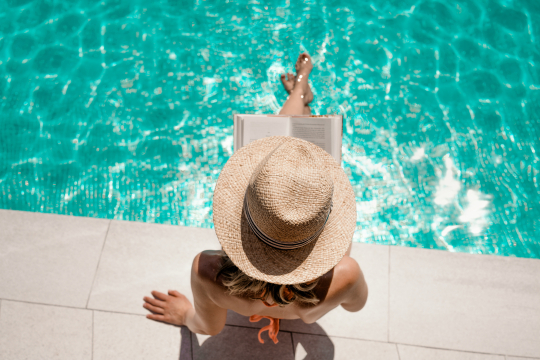 Frau mit Sonnenhut sitzt im Bikini an einem Pool Rand und liest ein Buch